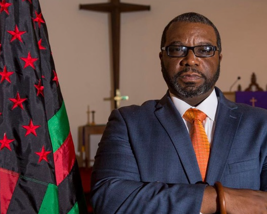 A man stands with his arms crossed next to a red, black, and green American flag. A cross hangs on the wall in the background.